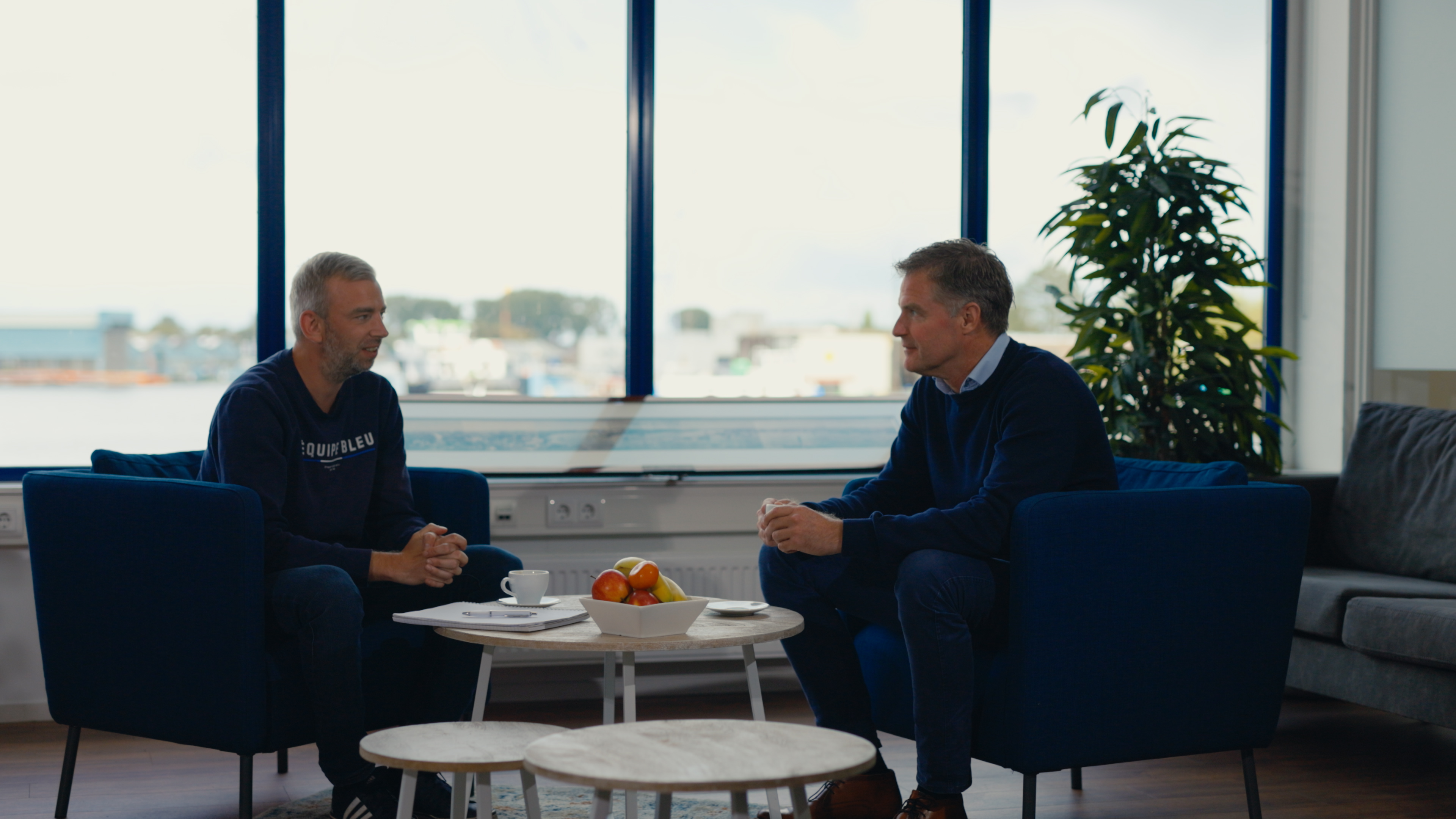 Two men are sitting at a table talking. You can look out onto a river.